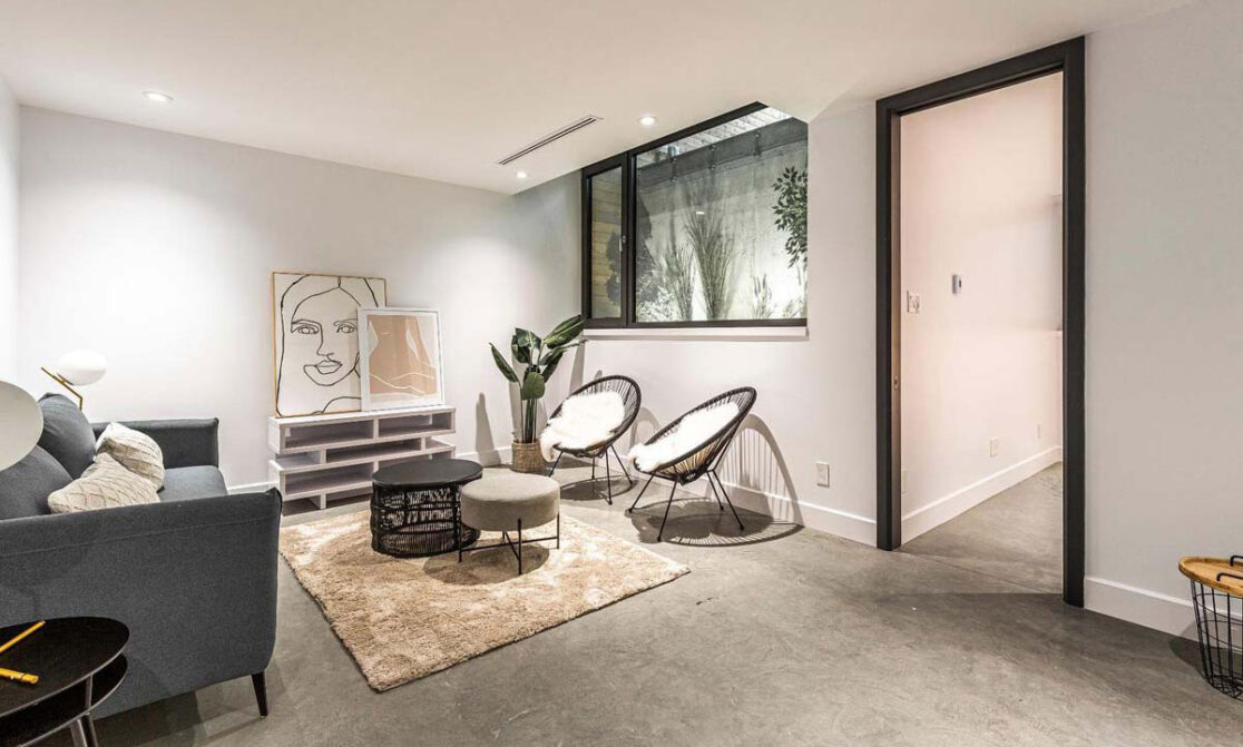 Sous-sol comprenant un espace salon avec un canapé, deux chaises et de grandes fenêtres agrémentées de plantes.