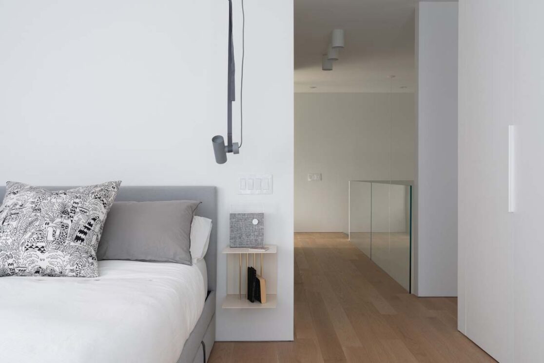 Chambre avec un lit gri et blanc avec vue sur le corridor menant vers les escaliers.